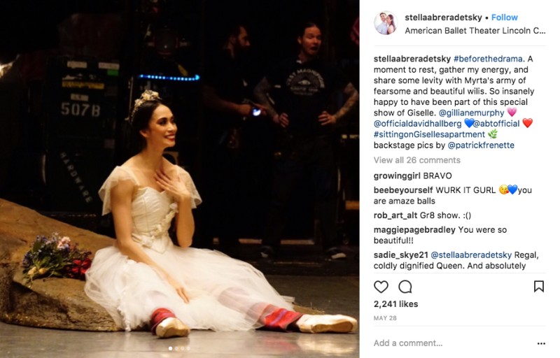 A behind-the-scenes image of American Ballet Theatre principal dancer Stella Abrera. She sits on a stone prop in a white romantic tutu. she wears knit warms up underneath.